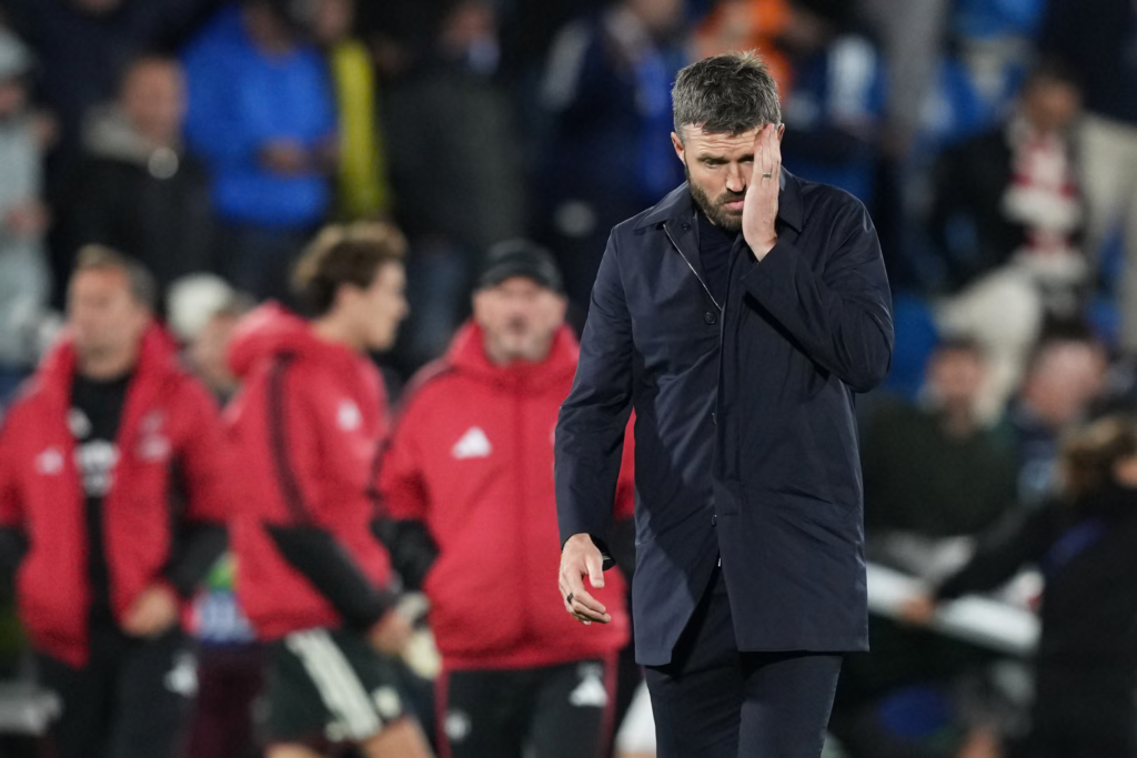 Manchester United’s Michael Carrick, in a dark navy coat with a zip at the collar and a wedding ring on his hand, presses his palm against his cheek and stares down with a pained frown. His grey hair and beard are short. Behind him, blurred United staff in red Adidas padded jackets stand beside a player in a black and white training kit, and Como fans in blue fill the stand.