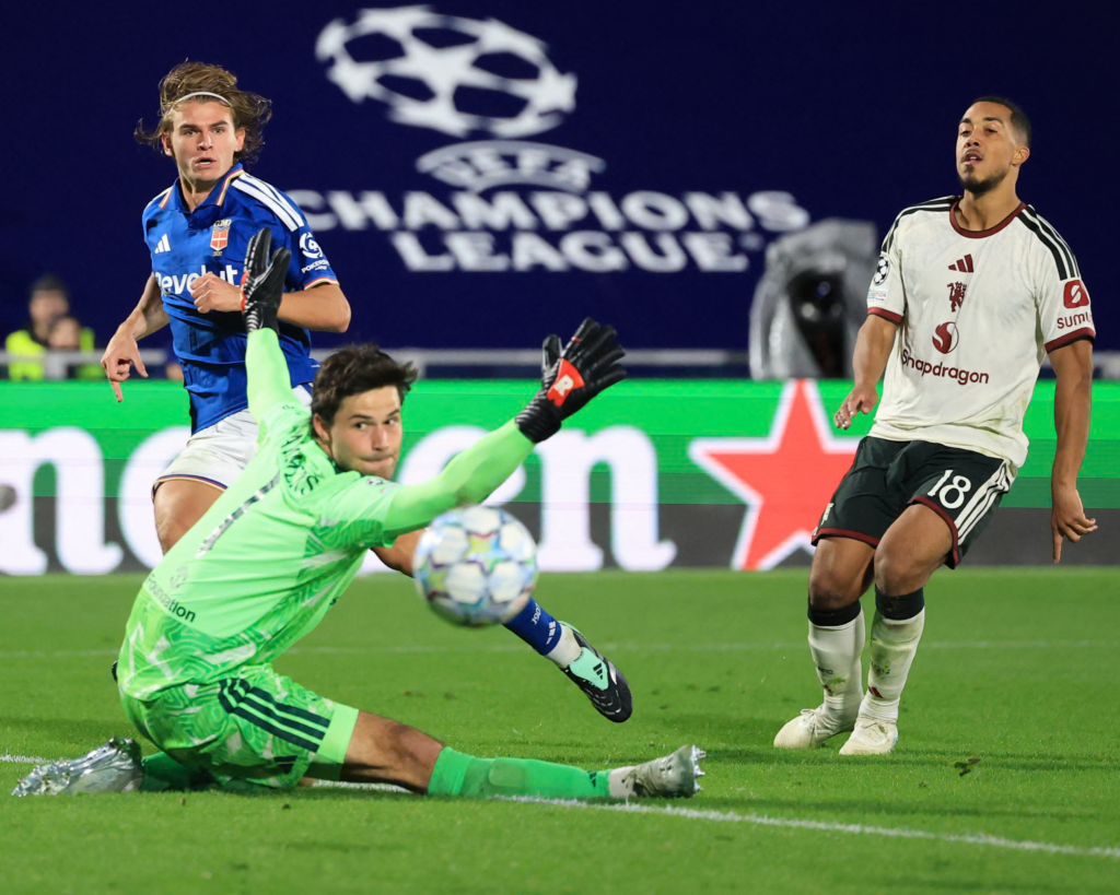 Manchester United goalkeeper Senne Lammens, in a bright green kit and black gloves, stretches out sideways along the grass with his arm raised as the ball flies past him. Como’s Nico Paz, in a blue kit with a white headband and long hair, stands behind him with his finger pointing down and his eyes on the ball. A United player wearing No. 18 in a white kit stands on the right watching. A Champions League banner and a green advertising board are behind them.