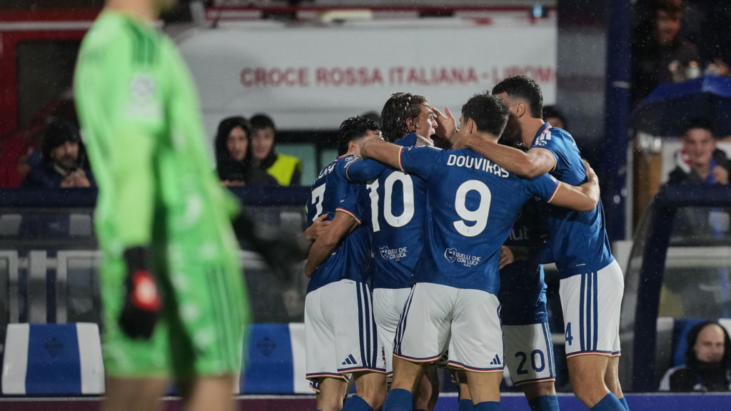 Como players in blue shirts and white shorts huddle together with their arms around each other in the rain. The shirts show No. 10, No. 7, No. 20 and No. 4, and “Douvikas” with No. 9 in the foreground. In the left foreground, a Manchester United goalkeeper in a bright green kit stands blurred with his head out of frame. An Italian Red Cross ambulance and fans in hoods are behind them.
