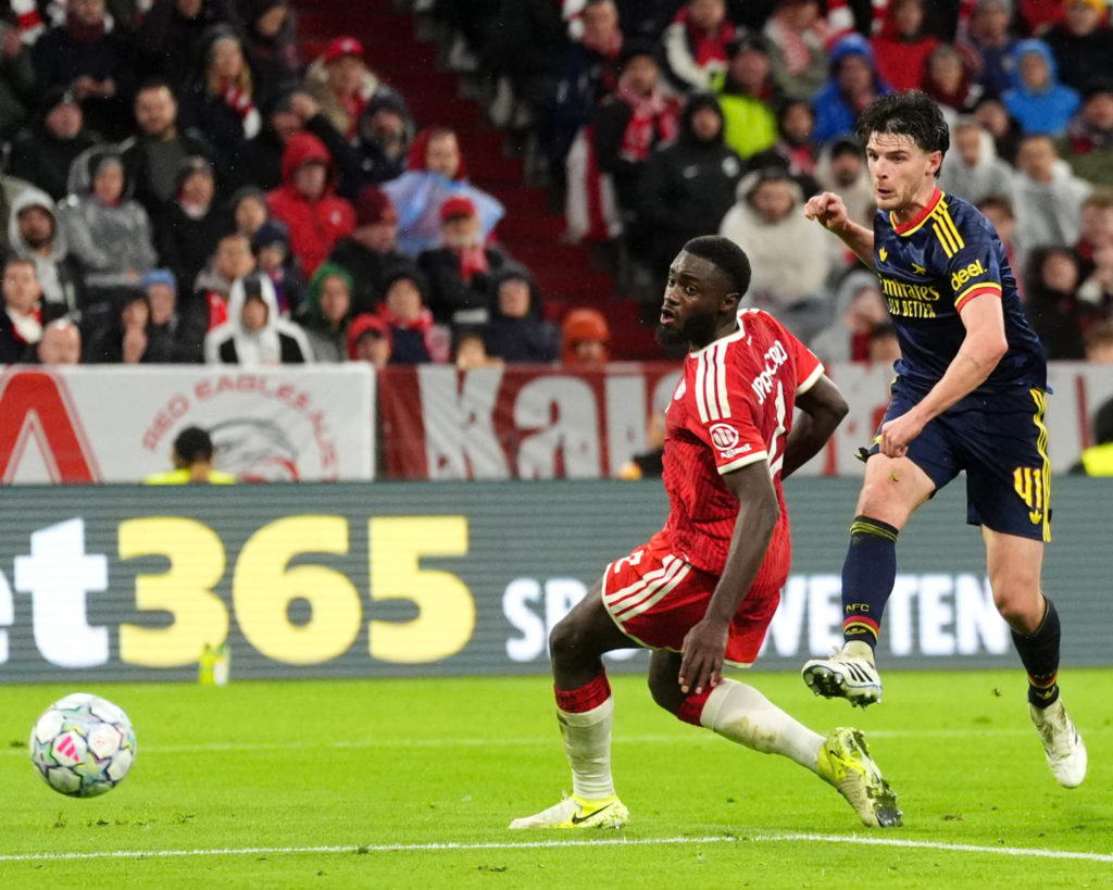 Arsenal’s Declan Rice, wearing No. 41 in a navy kit with yellow and red trim and the Emirates logo, follows through after striking the ball with his right foot, his fist clenched and his eyes ahead. A Bayern Munich player in a red kit with a beard stands bent forward beside him with a surprised look, one leg stretched out. The ball is on the grass at the bottom left, with a bet365 advertising board and a packed stand of fans in rain gear behind.