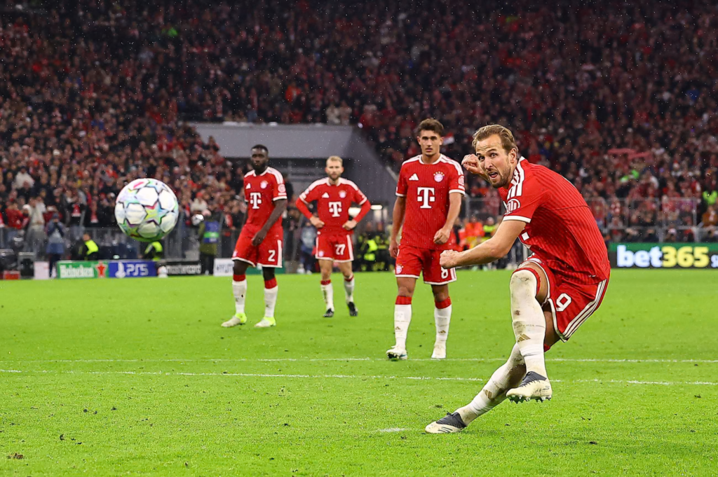 Bayern Munich’s Harry Kane, wearing No. 9 in a red kit, follows through with his right leg after striking a penalty, his eyes fixed ahead and his fist clenched. The ball is in the air at the left. Three Bayern teammates in red, wearing No. 2, No. 27 and No. 8, stand behind him watching, one with his hands on his hips. A packed stand and a bet365 advertising board are in the background.