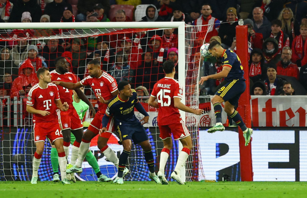 Arsenal’s Bruno Guimaraes, in a navy kit with yellow trim, leaps beside the goalpost on the right and heads the ball, with a No. 3 and a 9 partly visible on his shirt. Five Bayern Munich players in red defend in front of the goal, including No. 44 and “Pavlovic” No. 45, with a goalkeeper in green partly hidden behind them. An Arsenal teammate in navy with “Emirates Fly Better” on his chest watches from the centre. A packed stand of fans fills the background.