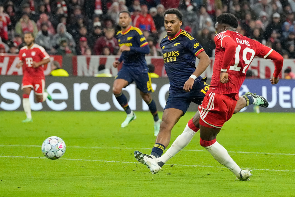 Bayern Munich’s Alphonso Davies, wearing No. 19 with “Davies” on the back of his red kit, swings his right leg through as he shoots, with his fists clenched. An Arsenal player in a navy kit with the Emirates logo and a white wristband runs alongside him, watching the ball. A second Arsenal player and a Bayern teammate wearing No. 7 are blurred behind. The ball is on the grass at the left, with a Mastercard advertising board and a packed stand behind.