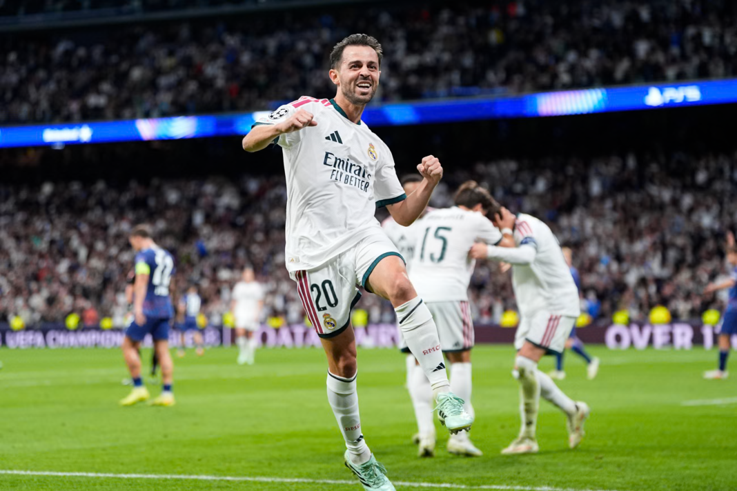 Real Madrid’s Bernardo Silva, wearing No. 20 in a white kit with the Emirates Fly Better logo, jumps with one knee raised and both fists clenched, smiling widely. Behind him, teammates in white, including one with No. 15, hug in celebration. A dejected Leipzig player in a blue kit with a captain’s armband stands on the left, with a packed Bernabéu stand and a PS5 advertising board in the background.