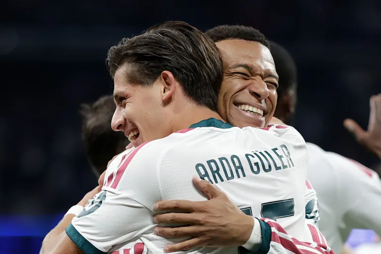 Real Madrid’s Arda Güler, in a white shirt with “Arda Güler” printed across the back and short dark hair, hugs a teammate and smiles with his eyes closed. The teammate, whose face is over Güler’s shoulder, grins widely with his eyes shut. Other players in white are partly visible behind them against a dark stadium background.