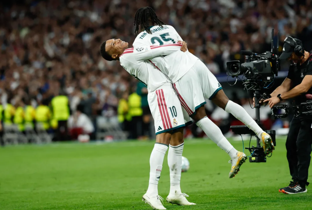 Real Madrid’s Yan Diomande, wearing No. 25 with his name printed on the back of his white shirt and his braids hanging down, is lifted off the ground in a hug by Kylian Mbappe, who wears No. 10 and tilts his head back, smiling. Diomande’s legs are raised behind him. A TV cameraman with a large camera stands on the right, with stewards in yellow vests and a packed stand behind.