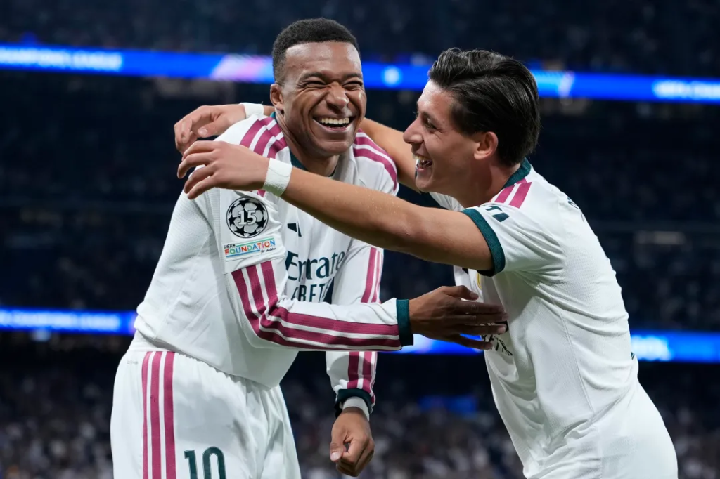 Two Real Madrid players in white kits with pink and green trim embrace and laugh. On the left, Kylian Mbappe, wearing No. 10 and a Champions League badge on his sleeve, grins with his eyes closed. On the right, Arda Güler, with dark slicked-back hair and a white wristband, smiles with his arm around him. A dark stadium and a blue Champions League advertising board are blurred behind them.
