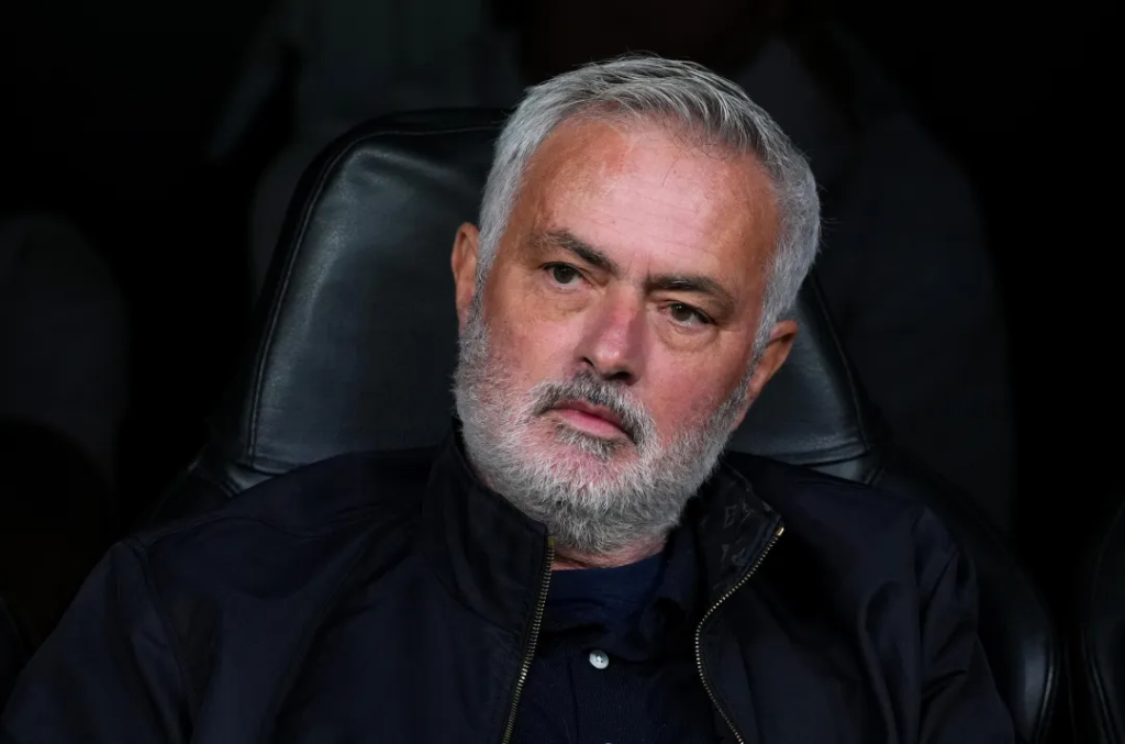 Real Madrid coach Jose Mourinho, with short grey hair and a grey beard, sits in a black leather seat wearing a dark zip-up jacket over a navy shirt. He looks off to the side with a serious, slightly furrowed expression against a dark background.