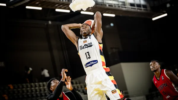 Nam Blazers’ Fataki Nyembo, wearing No. 13 in a white “Blazers” jersey with the Crane Millers logo and the Uganda flag, rises above the rim with the ball in his right hand, ready to dunk. A net hangs above him. Two defenders in red and black kits are around him, one with arms raised at the bottom left and one at the right, with empty seats behind.