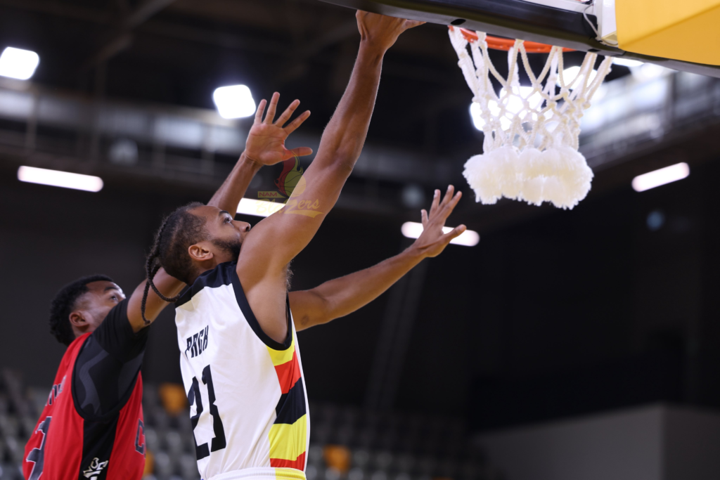 Nam Blazers’ No. 21, in a white jersey with the Uganda flag colours down the side and braided hair, stretches his right arm up to lay the ball in at the rim, with a white net hanging below. A defender in a red and black kit reaches up behind him with one arm raised.
