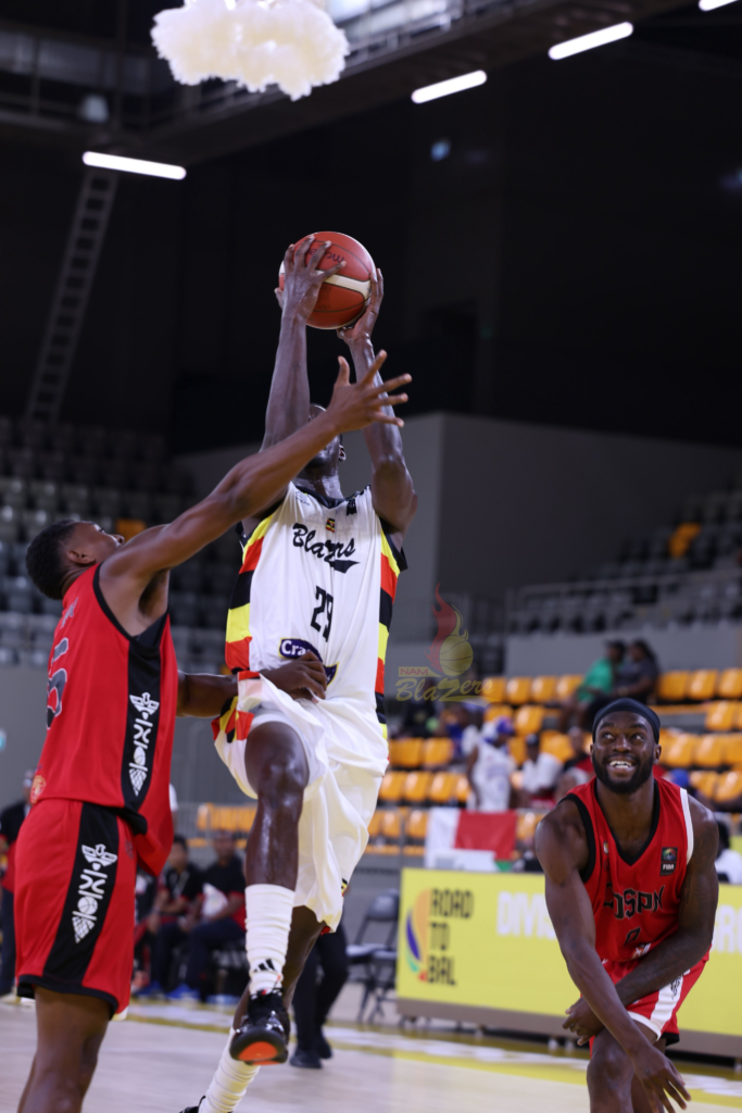 Nam Blazers’ No. 29, in a white “Blazers” jersey with the Uganda flag colours, rises off the floor holding the ball above his head as a defender in a red and black kit reaches across him on the left. A second defender in red with a black headband crouches at the bottom right, smiling and looking up. A “Road to BAL” board and yellow seats are behind them.