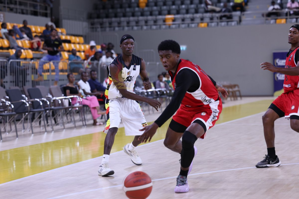A Nam Blazers player in a white jersey with the Uganda flag colours and a black headband stands with his arms out, guarding a player in a red and black kit who drives past with his eyes on the ball. The ball is bouncing on the court at the bottom, a second player in red is at the right edge, and spectators sit in yellow and grey seats behind.